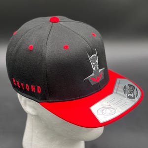 May include: Black and red baseball cap with a black and grey embroidered Batman logo on the front. The cap has a red underside and the word "BEYOND" embroidered in red on the side.