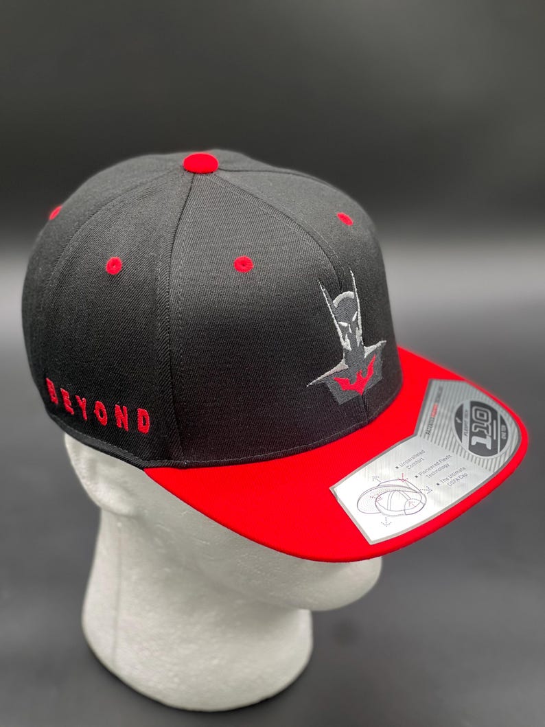 May include: Black and red baseball cap with a black and grey embroidered Batman logo on the front. The cap has a red underside and the word "BEYOND" embroidered in red on the side.