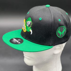 May include: Black and green baseball cap with a flat brim. The cap features an embroidered design on the front and side, with a green bill and top button. The brand name "DECKY" is visible on the bill.
