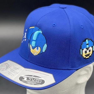 May include: Royal blue Flexfit baseball cap featuring embroidered Mega Man character designs. The cap has a curved brim and a Flexfit 110 tag. The Mega Man character is on the side and front of the cap.