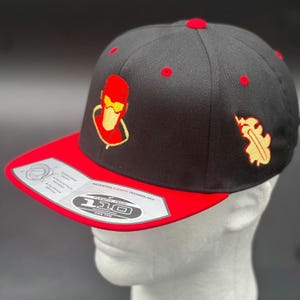 May include: A black baseball cap with a red brim and red eyelets. The front features an embroidered red and yellow character graphic. A yellow flame-like emblem is on the side. A silver sticker on the brim reads Flexfit Tech 110 One Ten.