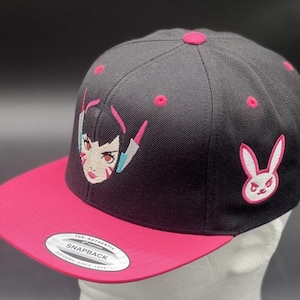 May include: Black and pink snapback hat featuring embroidered artwork. The hat has a pink bill and pink accents on the crown. The front of the hat displays a character's face, and the side features a white and pink bunny logo.