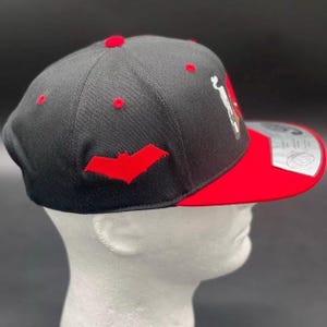 May include: Black and red baseball cap with a flat brim. The cap features a red bat logo on the side and red accents on the top. The brim is red, and the cap is displayed on a white mannequin head.