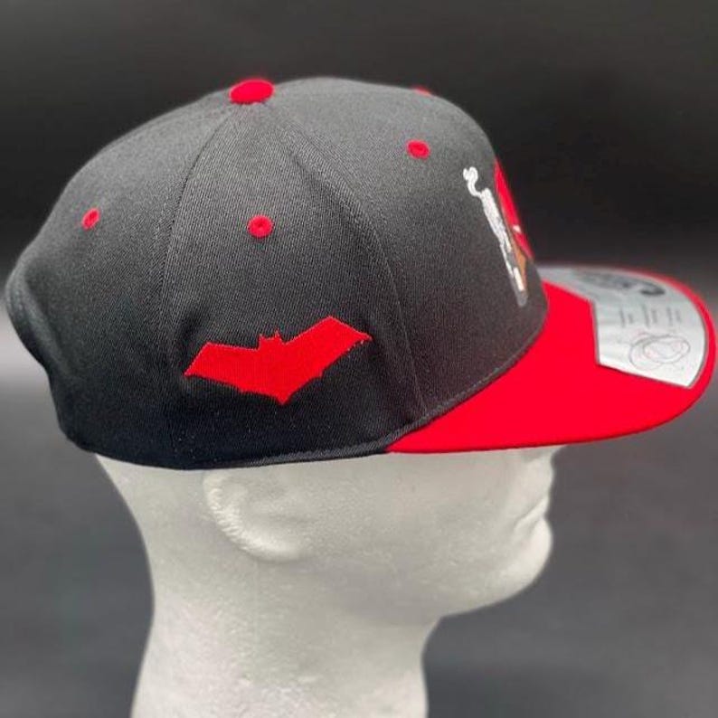 May include: Black and red baseball cap with a flat brim. The cap features a red bat logo on the side and red accents on the top. The brim is red, and the cap is displayed on a white mannequin head.