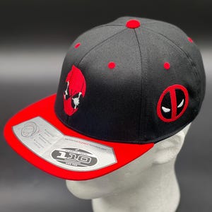 May include: Black and red baseball cap featuring the Deadpool logo. The cap has a red brim and red embroidered details, including the Deadpool mask and logo. The cap is displayed on a white mannequin head.