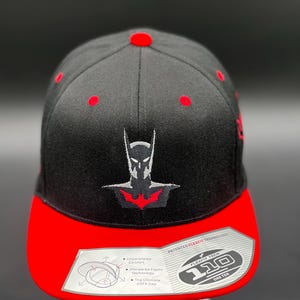May include: Black and red baseball cap with a black embroidered Batman logo. The cap features Flexfit technology and the text "110 One Ten".