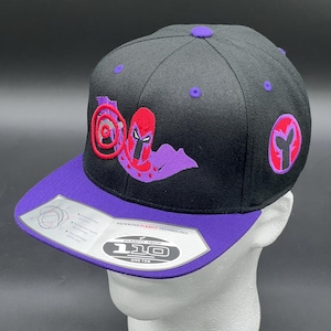 May include: Black and purple baseball cap with a red and purple embroidered design of a superhero with a red helmet and a purple cape. The cap has a purple brim and a black crown. The cap has a white label with the text "Flexfit Tech 110 One Ten" and "Patented Flexfit Technology".