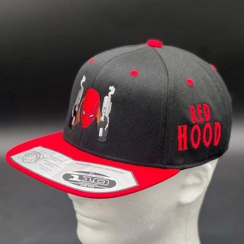 May include: Black and red baseball cap with the text "RED HOOD" embroidered in red. The cap features an embroidered design of a red character with guns. The brim is red, and the cap is on a white mannequin head.