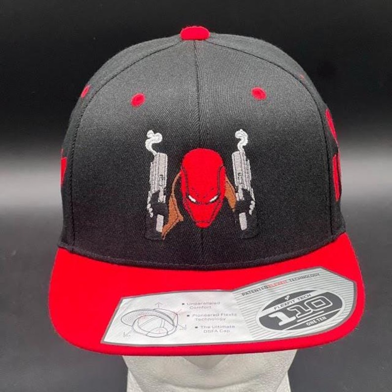 May include: Black and red baseball cap featuring an embroidered design of a character with a red mask and two guns. The cap has a red brim and a Flexfit Tech label.