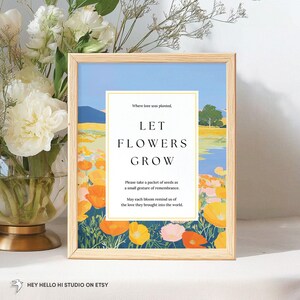 May include: A framed art print with a landscape scene featuring a field of orange and yellow flowers, a blue sky, and the text "LET FLOWERS GROW". The frame is made of light-colored wood.