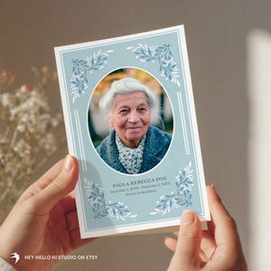 May include: A memorial card held in hands, featuring a portrait of an elderly woman within a light blue frame. The card includes floral accents and the text "PAULA REBECCA FOX" with dates and "Forever in Our Hearts". The Etsy shop name is visible.