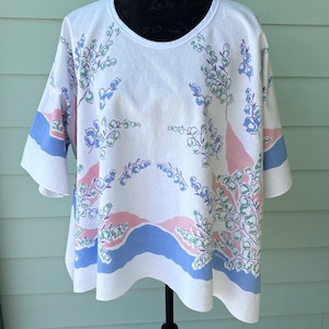 May include: White, short-sleeved top with a scoop neck, featuring a floral pattern in shades of blue, green, and pink. The sleeves have blue and pink stripes. The top is displayed on a black mannequin.
