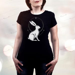 May include: Black t-shirt featuring a detailed illustration of a white rabbit within a circular design. The rabbit is depicted in a seated position, with long ears and a serene expression. The design is centered on the shirt, creating a striking visual.