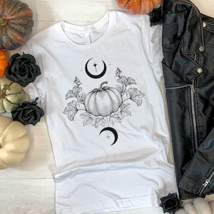 May include: White t-shirt with a black and white pumpkin graphic, surrounded by leaves and crescent moons. The image also includes a black leather jacket, pumpkins in various colors, and black roses. A seasonal, fall-themed design.