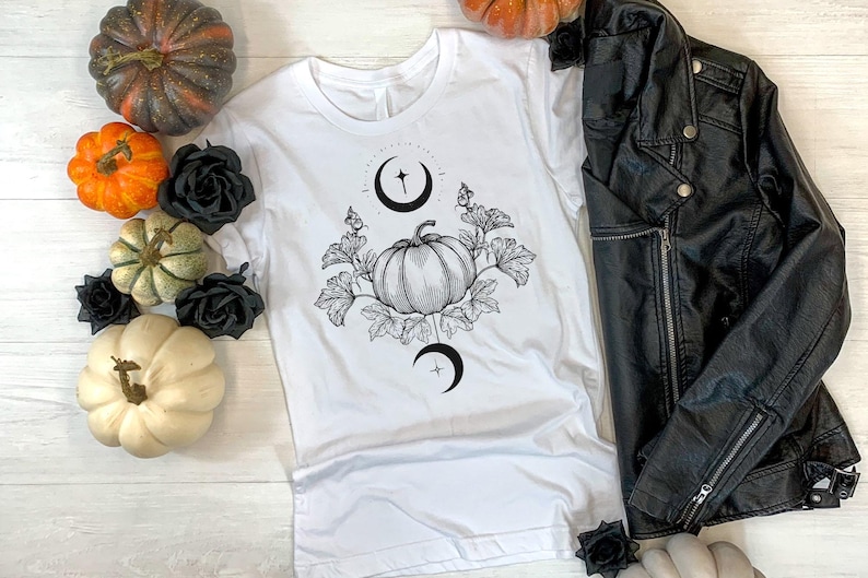 May include: White t-shirt with a black and white pumpkin graphic, surrounded by leaves and crescent moons. The image also includes a black leather jacket, pumpkins in various colors, and black roses. A seasonal, fall-themed design.