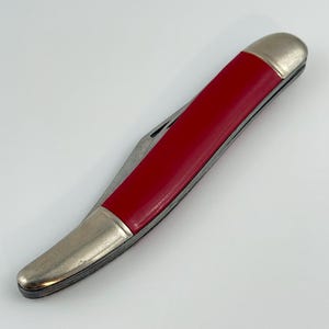 May include: A closed pocket knife with a bright red handle and silver-colored metal accents. The knife has a curved shape and appears to be made of durable materials. The blade is partially visible.