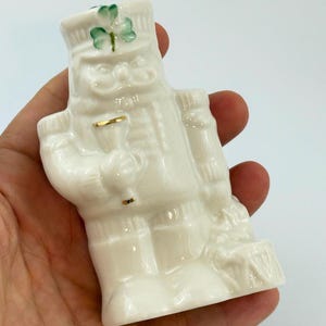 May include: A white ceramic figurine of a soldier, possibly a nutcracker, with a green shamrock on the hat. The figurine has gold accents and is holding a small object. The figure is in a standing position.