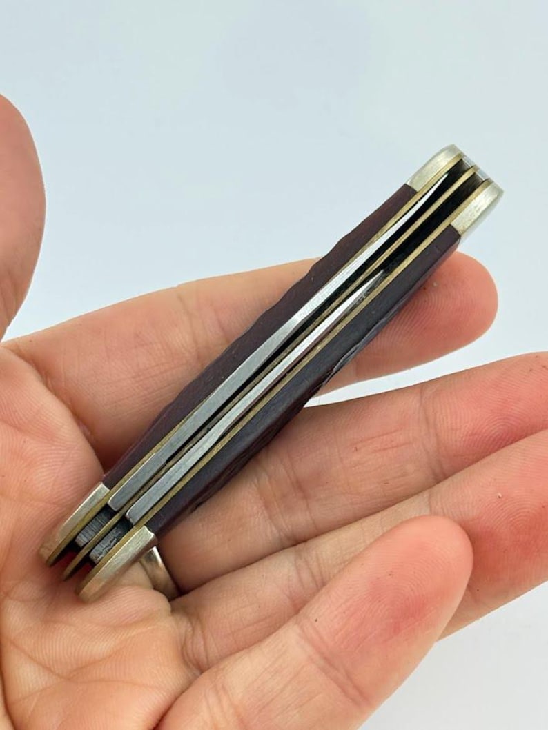 Może przedstawiać: Zamknięty scyzoryk z ciemnobordową rączką i srebrnymi metalowymi akcentami. N&oacute;ż ma wiele ostrzy i klasyczny design. N&oacute;ż jest trzymany w dłoni.