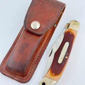 May include: A vintage Old Timer pocket knife with a brown handle and a stainless steel blade, next to a matching leather sheath. The sheath has a snap closure and the words "Old Timer" embossed on it.