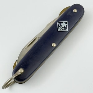 May include: A navy blue pocket knife with a silver blade and a metal loop. The knife has a Cub Scouts BSA emblem on the side. The knife is closed, with the blade tucked inside the handle.