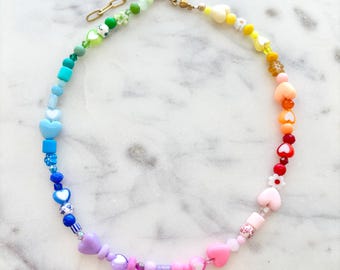 May include: A rainbow-colored beaded necklace with white heart-shaped charms. The necklace is made with a variety of beads, including squares, circles, and flowers.