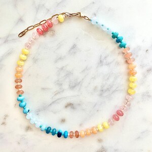 May include: A rainbow-colored beaded necklace with a gold clasp. The beads are made of different types of stones, including turquoise, aquamarine, and rose quartz.