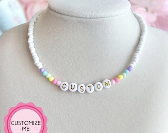 May include: A white beaded necklace with a rainbow of colored beads and gold letters spelling "CUSTOM" hanging on a white mannequin.
