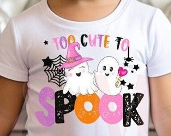 Puede incluir: Una niña pequeña con una camiseta blanca con la frase "Too Cute to Spook" y un tutú morado. La camiseta presenta un gráfico de dos fantasmas, un sombrero de bruja y una telaraña. Un lazo morado está en el pelo de la niña. El fondo incluye calabazas decorativas.