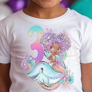 Pode incluir: Uma menina pequena vestindo uma camiseta branca com um gráfico colorido de uma sereia montando um golfinho e o número "3". O gráfico tem cores roxo, azul e verde. A menina tem um coque no cabelo e está sorrindo.