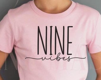 Könnte beinhalten: Hellrosa T-Shirt mit dem schwarzen Schriftzug "NINE vibes". Das kurzärmelige Oberteil wird von einem jungen Mädchen mit blonden Haaren im Dutt getragen. Der Hintergrund ist einfarbig grau.