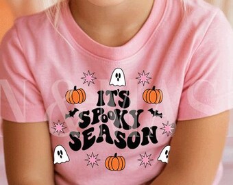 Puede incluir: Una niña pequeña con una camiseta rosa con las palabras "It's Spooky Season" en blanco y negro, junto con calabazas, murciélagos y fantasmas naranjas. La niña está sentada frente a un fondo de calabazas.