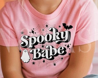 Puede incluir: Una niña pequeña lleva una camiseta rosa con las palabras "Spooky Babe" en un diseño en blanco y negro. La camiseta presenta un fantasma, telarañas y un murciélago. El fondo incluye calabazas.