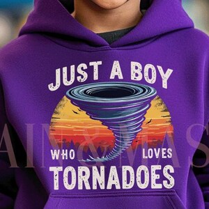 Op de afbeelding: Een paarse hoodie met de tekst "JUST A BOY WHO LOVES TORNADOES" en een afbeelding van een tornado boven een zonsondergang. De hoodie wordt gedragen door een lachend kind.