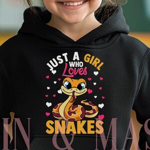 Könnte beinhalten: Schwarzer Kapuzenpullover mit dem Text "Just a Girl Who Loves Snakes" und einer Cartoon-Schlangenillustration. Das Design zeigt eine lächelnde Schlange mit Herzakzenten. Der Kapuzenpullover wird von einem jungen Mädchen getragen.