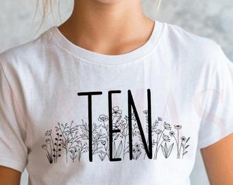 Könnte beinhalten: Ein weißes kurzärmeliges T-Shirt mit dem Wort TEN in fetter schwarzer Schrift. Die Buchstaben sind in eine feine schwarze Strichzeichnung aus verschiedenen Wildblumen und Stängeln integriert, die sich über den unteren Teil des Motivs erstreckt.