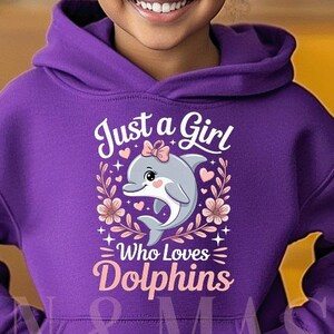 Puede incluir: Una niña pequeña lleva una sudadera con capucha morada con el texto "Just a Girl Who Loves Dolphins". La sudadera con capucha presenta un gráfico de un delfín con detalles florales. La niña está sonriendo.