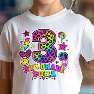 May include: A white short-sleeved t-shirt featuring a colorful graphic with a large number 3 in a rainbow leopard print. The design includes icons like a lightning bolt, star, heart, peace sign, and lollipop, with the text 3RD GRADE CORA.