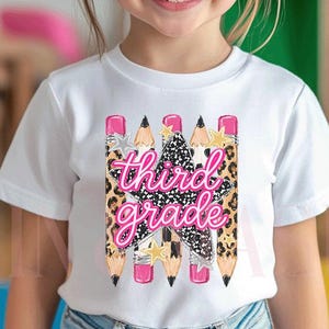 Könnte beinhalten: Ein weißes kurzärmeliges T-Shirt mit einem Grafikdesign aus vier vertikal angeordneten Bleistiften und einem zentralen Stern. Der Text "third grade" ist in einer rosa Schreibschrift über den Stern gedruckt. Die Stifte haben Leopardenmuster.