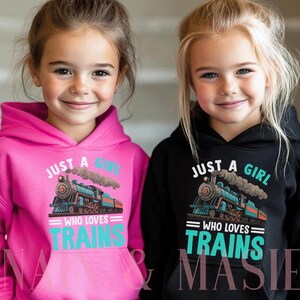 Puede incluir: Dos niños con sudaderas con capucha. Una es rosa intenso, la otra negra. Ambas sudaderas tienen un gráfico de tren con el texto "JUST A GIRL WHO LOVES TRAINS". El tren es azul, rojo y gris.