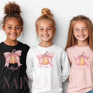 May include: Three girls wearing long-sleeved shirts with a pumpkin design and a pink bow. The shirts are black, white, and pink. Each shirt has a name printed below the pumpkin: Abigail, Emma, and Gigi. The shirts are for children.
