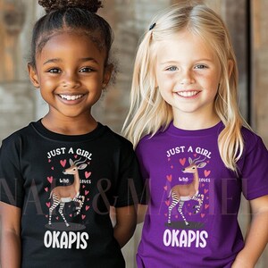 Op de afbeelding: Twee jonge meisjes dragen zwarte en paarse t-shirts met de tekst "JUST A GIRL who loves OKAPIS". De shirts hebben een afbeelding van een okapi met harten.