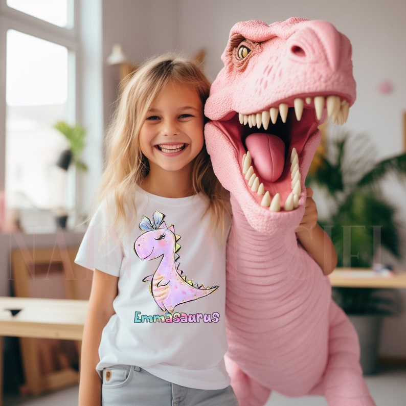 May include: A white short-sleeved t-shirt featuring a graphic of a pink cartoon dinosaur with a blue bow on its head. The text "Emmasaurus" is printed below the dinosaur in colorful letters. The shirt is paired with light gray denim pants.
