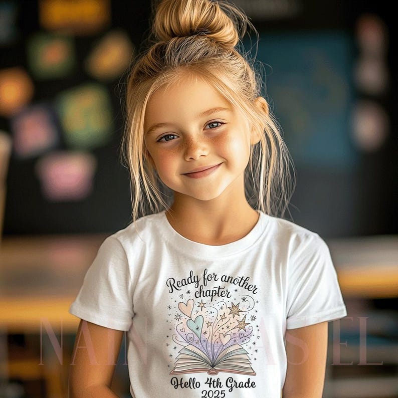 Puede incluir: Una ni&ntilde;a rubia con el pelo recogido en un mo&ntilde;o sonr&iacute;e, vistiendo una camiseta blanca. La camiseta presenta un gr&aacute;fico colorido de un libro abierto con corazones, estrellas y el texto "Ready for another chapter" y "Hello 4th Grade 2025".