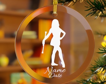 May include: A clear, circular glass ornament featuring a white silhouette of a baton twirler holding a baton. Below the figure, the text reads Name and Date in a white script font. The ornament hangs from a gold ribbon on a tree branch.