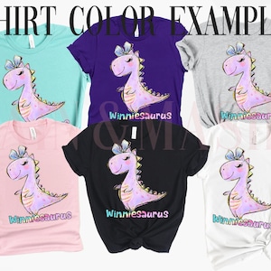 May include: Six t-shirts in light blue, purple, gray, light pink, black, and white. Each shirt features a graphic of a pink dinosaur with a blue bow and the text Winniesaurus. The image is titled SHIRT COLOR EXAMPLES at the top in black text.