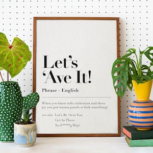 Può includere: Una stampa incorniciata con sfondo bianco e testo nero che recita "Let's 'Ave It!" Il testo è una definizione della frase, che è descritta come un'espressione inglese. La stampa è incorniciata in una cornice di legno marrone chiaro e si trova su una superficie bianca con puntini neri.