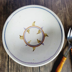 Puede incluir: Un cuenco de cerámica blanca con un borde azul y morado. El cuenco tiene un diseño marrón y morado en el centro, que se asemeja a un sol con rayos.