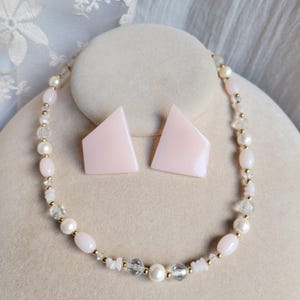 Könnte beinhalten: Ein Schmuckset bestehend aus einer Halskette und zwei Ohrringen. Die Kette besteht aus abwechselnden hellrosa ovalen Perlen, weißen Perlen, klaren facettierten Perlen und kleinen goldfarbenen Zwischenperlen. Zwei passende hellrosa Ohrringe.