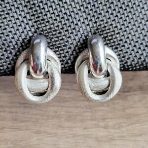 May include: A pair of silver tone knot-style earrings. The earrings are made of a smooth metal with a textured finish.