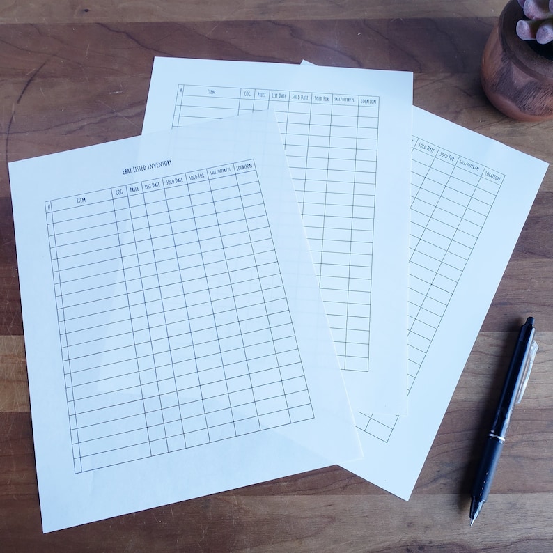 May include: Several sheets of white paper with a grid layout, labeled "Etsy Listed Inventory." A black pen rests on a wooden surface next to the papers. The papers are designed for tracking item details.